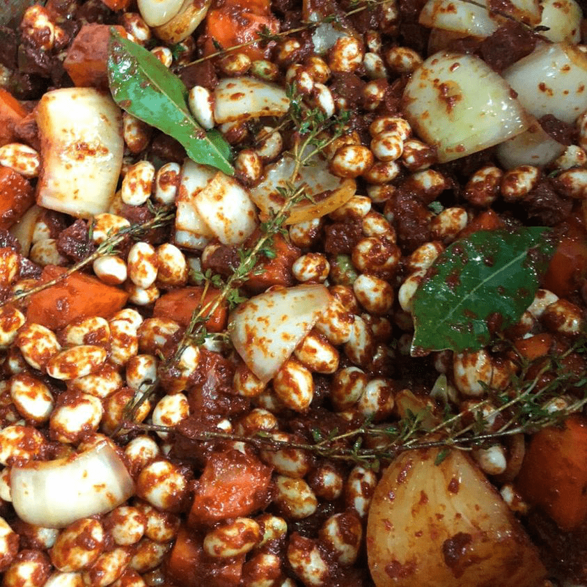 A close-up of a rich, hearty stew from Corner House, featuring white beans, chunks of meat, carrots, onions, bay leaves, and fresh thyme