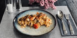 Eat At My Kitchen's elegant seafood pasta dish, featuring tagliatelle with shrimp, scallops, and cherry tomatoes, presented on a textured placemat with a sprig of flowers in the background.