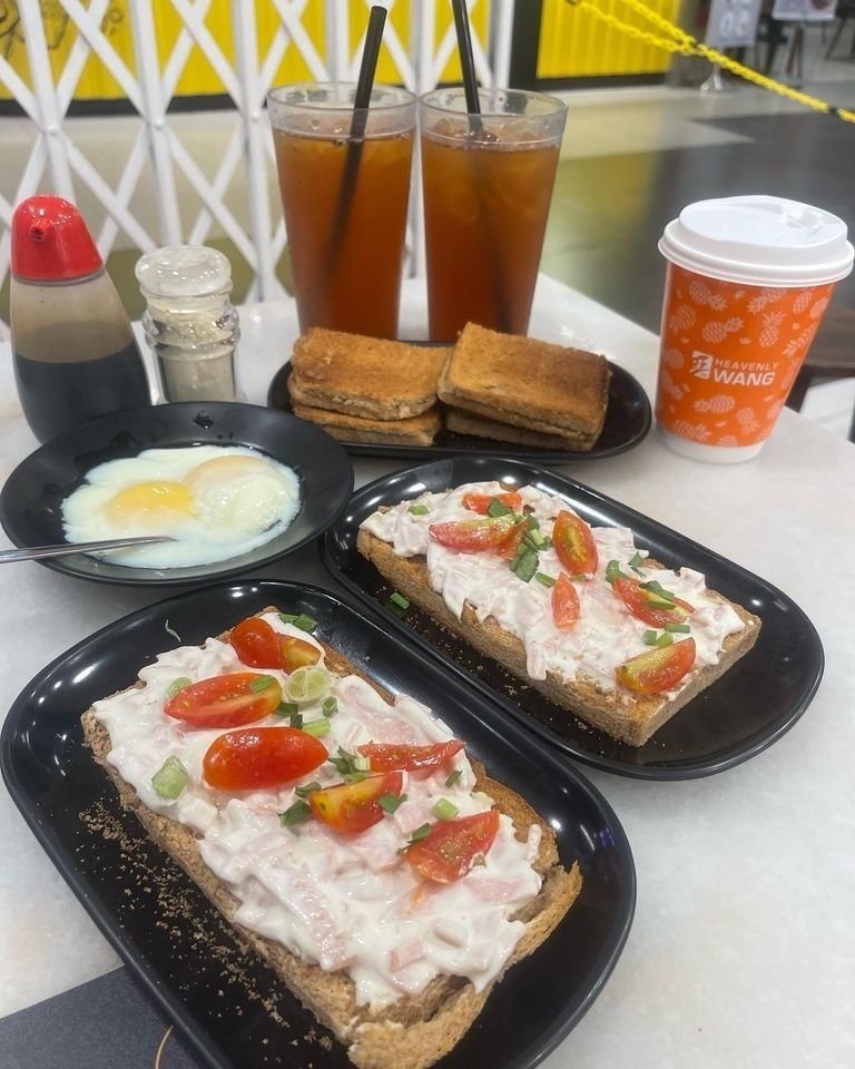 A breakfast set from Heavenly Wang Cafe, featuring open-faced toasts with creamy topping and tomatoes, kaya toast, soft-boiled eggs, two iced drinks, and a takeaway coffee cup