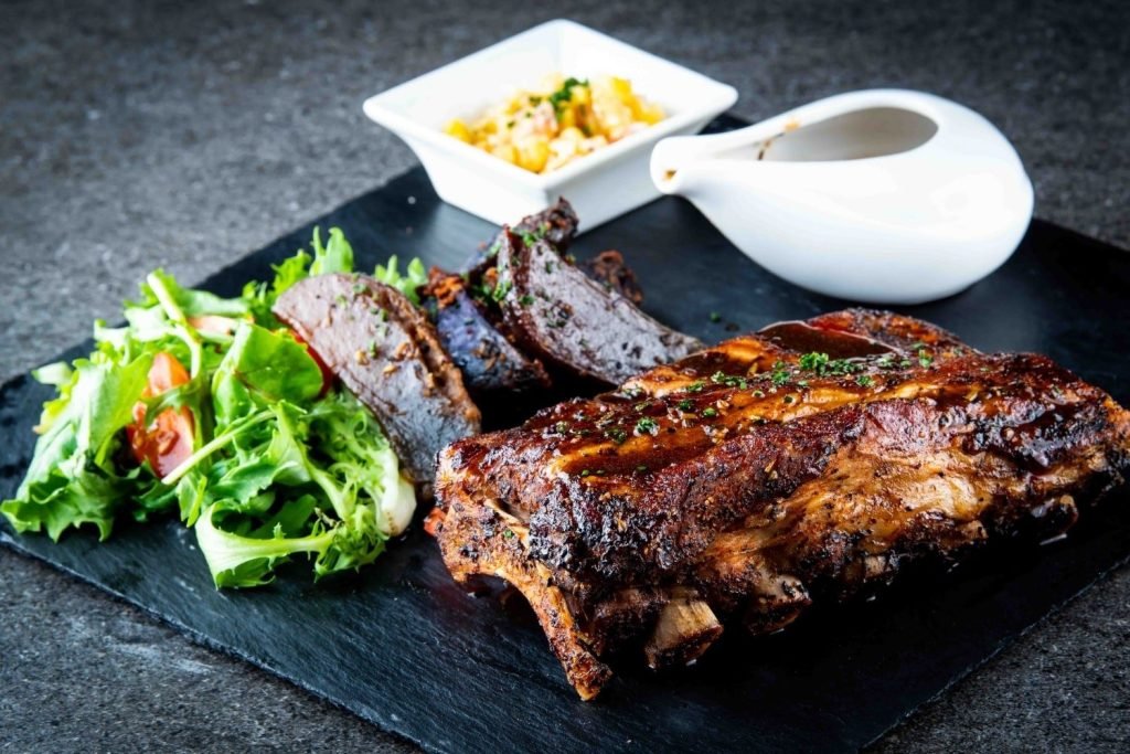 A glistening rack of BBQ pork ribs from Hopscotch, served on a black slate platter.