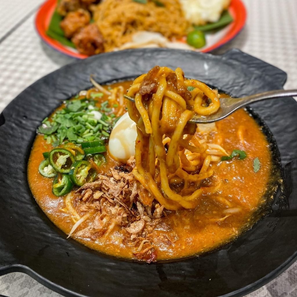 A fork lifts a serving of thick yellow noodles coated in rich, savory gravy from a black bowl of Mee Rebus at My Makan Place.