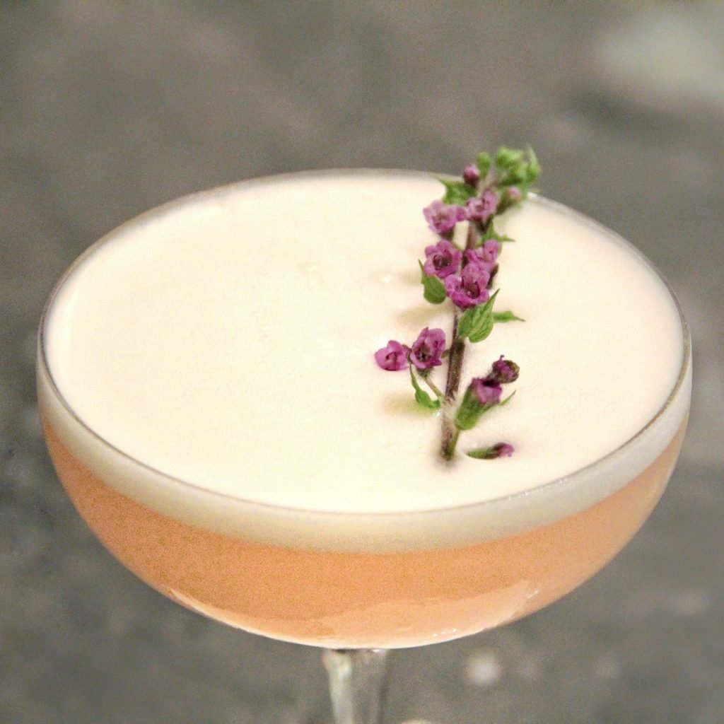 A pink cocktail in a coupe glass with white foam and a purple flower garnish