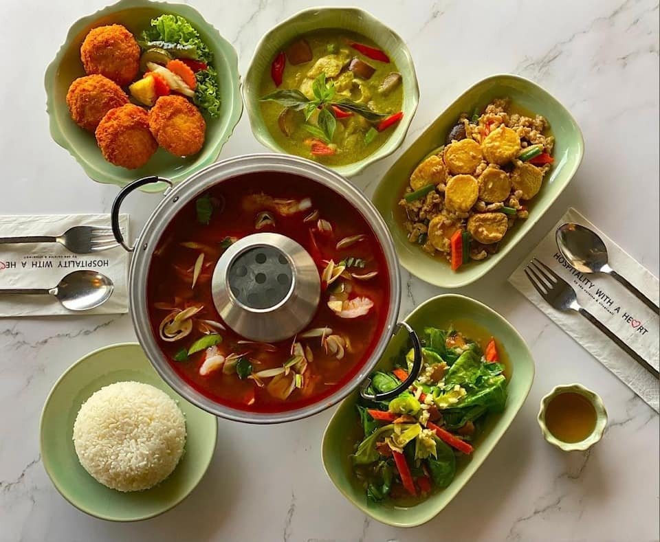 A flat lay of a Thai meal from Thai-Accent, featuring Tom Yum soup in a hot pot, green curry, stir-fried tofu, fried fish/prawn cakes, stir-fried vegetables, and rice, on a marble table.