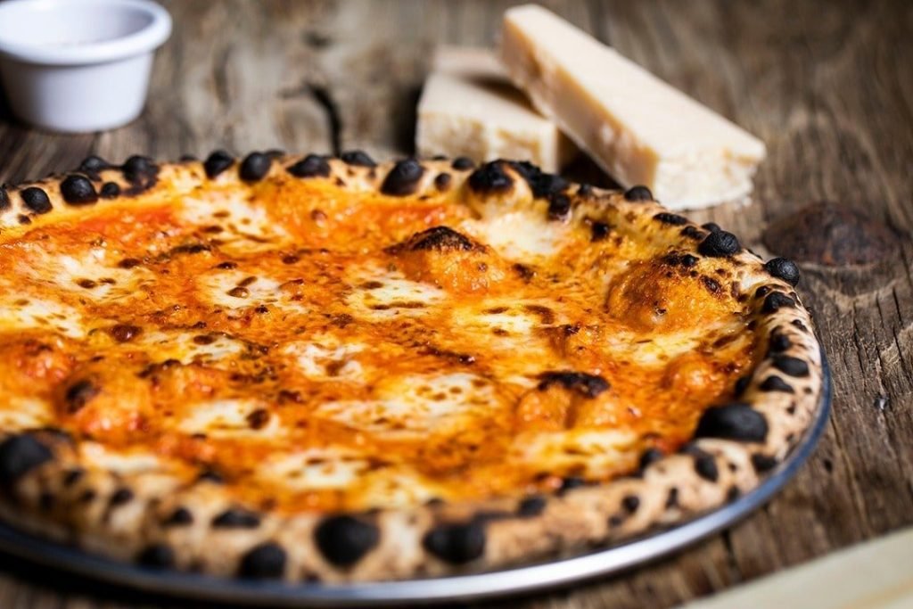 The signature pizza from Tong Dim Noodle Bar, featuring a blistered leopard-spot crust, served on a rustic wooden table with a side of parmesan cheese