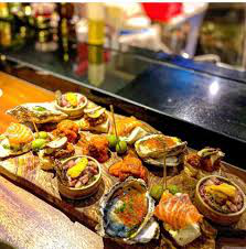 A long wooden board laden with an assortment of gourmet seafood appetizers, including oysters (some baked/topped), slices of salmon, small bowls with savory mixtures