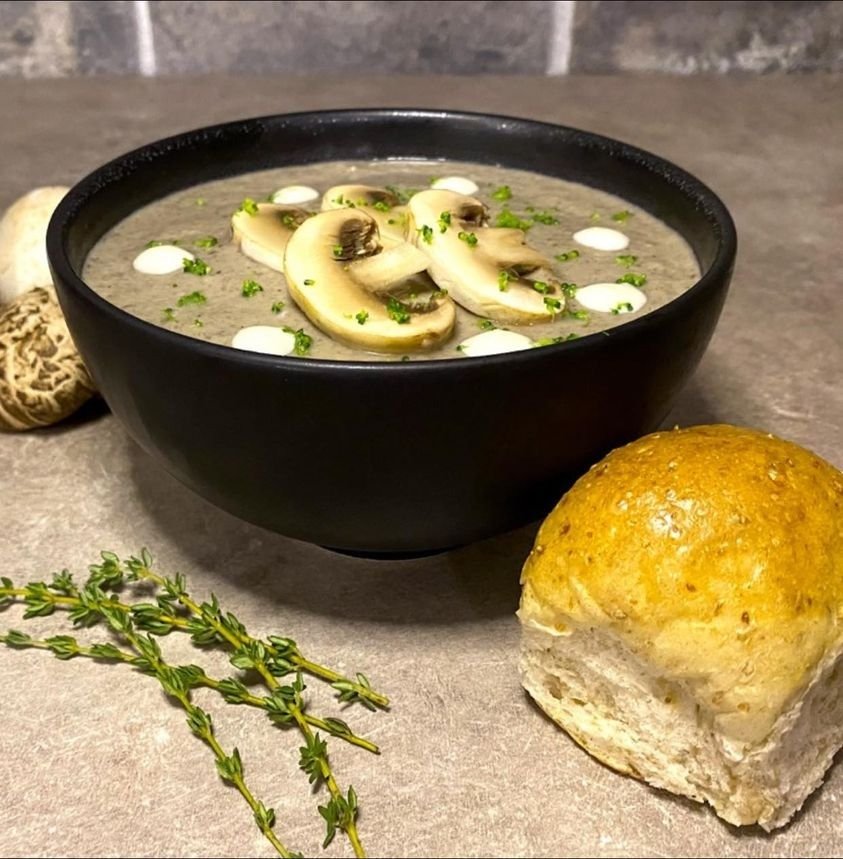 A bowl of creamy mushroom soup from Attap House, served in a black bowl, topped with fresh mushroom slices, dollops of cream, and chopped herbs, accompanied by a golden-brown bread roll and sprigs of fresh thyme.