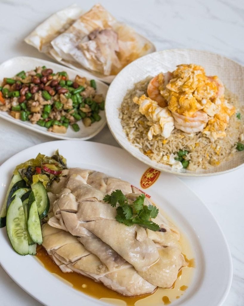 A delicious spread of classic Singaporean dishes from Boon Tong Kee, featuring their signature Hainanese poached chicken with cucumber, prawn fried rice topped with scrambled egg, stir-fried green beans with minced meat, and paper-wrapped chicken.