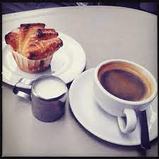 Cup of black coffee, a flaky pastry, and a small jug of milk on a light-colored cafe table.