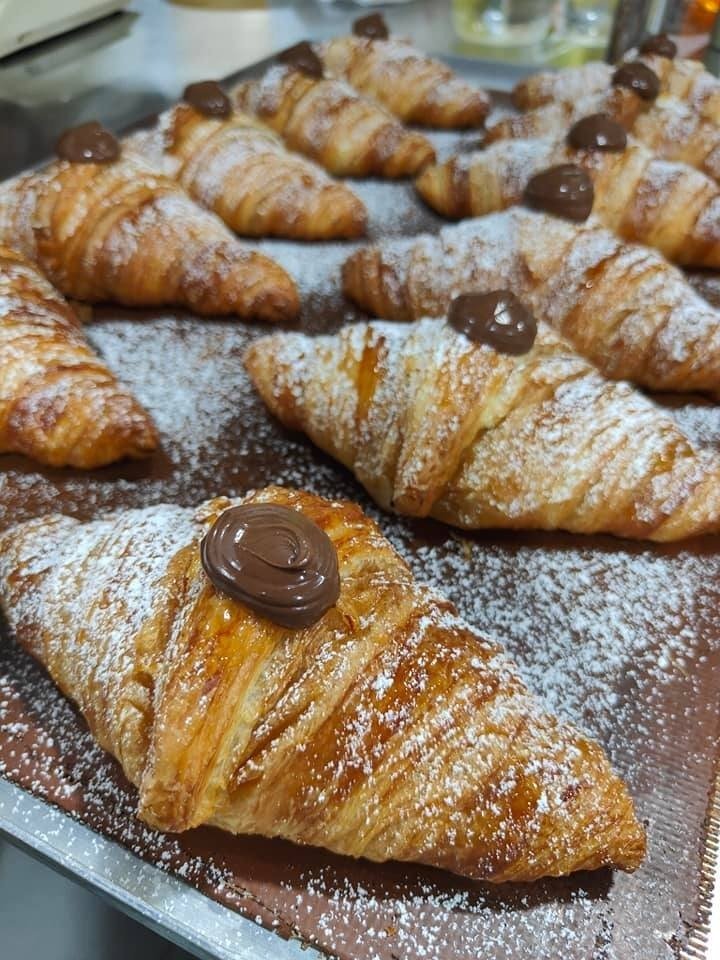A tray of freshly baked golden croissants from Brunetti, each topped with a dollop of chocolate spread and generously dusted with powdered sugar.