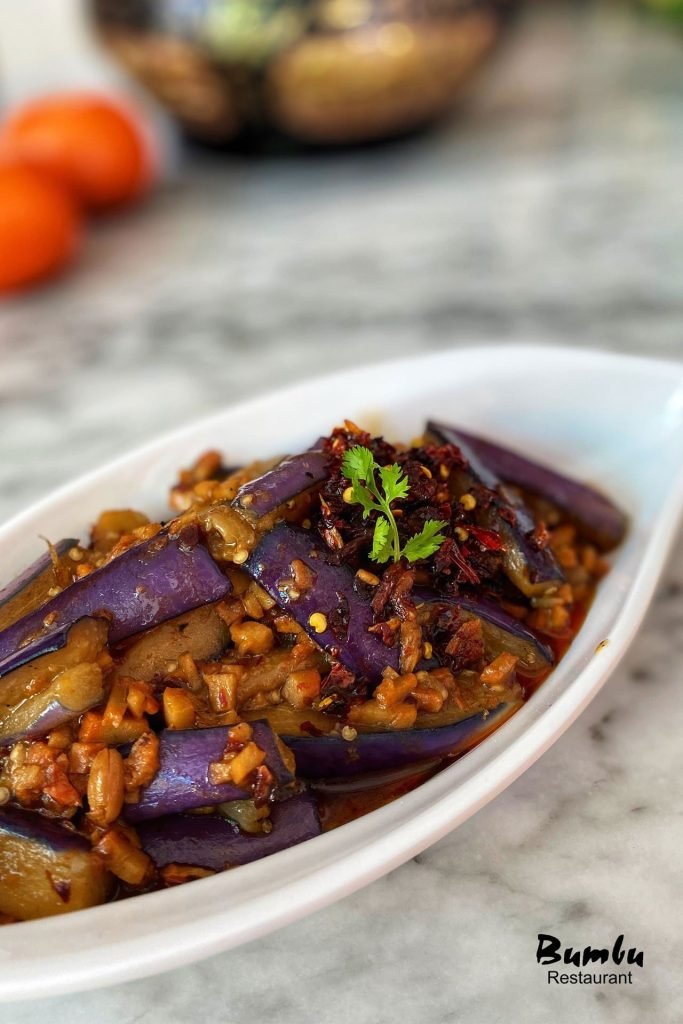 A dish of spicy stir-fried eggplant from Bumbu Restaurant, featuring sliced purple eggplant in a savory sauce with minced ingredients and chili flakes, garnished with cilantro, served in a white oval dish on a marble tabletop.