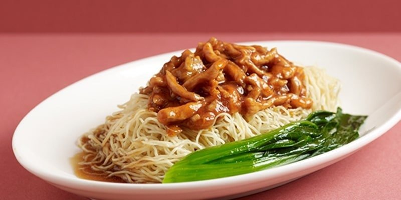 A dish from Canton Paradise featuring thin egg noodles topped with savory shredded pork in a brown sauce, served with a side of blanched green leafy vegetables (likely choy sum) on a white oval plate against a plain reddish-pink background