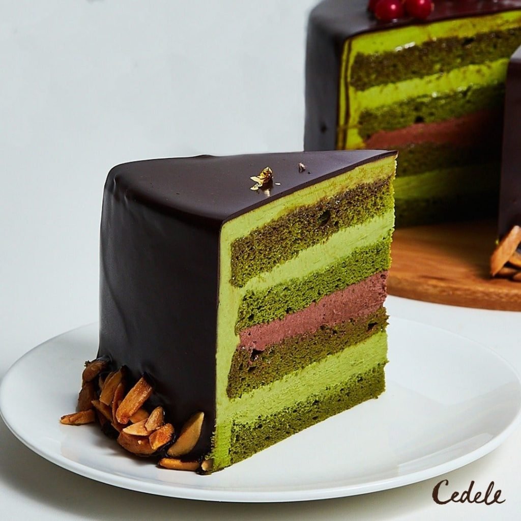 A slice of Cedele Bakery Kitchen's multi-layered cake on a white plate, featuring alternating green sponge and light green cream layers with a central chocolate cream layer, all enrobed in a glossy dark chocolate ganache.