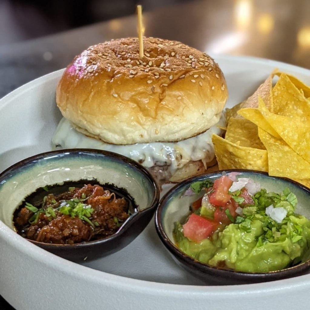 A burger from Chimichanga, featuring a sesame seed bun and melted cheese, served on a light-colored plate with tortilla chips, a small bowl of guacamole topped with pico de gallo, and a small bowl of red salsa.