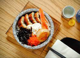 Japanese unagi donburi (eel rice bowl) in a stone bowl, topped with crab stick, egg, seaweed, and fish roe, served on a wooden board.