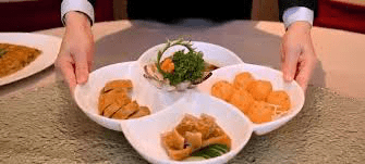 Server's hands in a suit presenting a white, four-leaf clover shaped serving platter containing four different Asian appetizers.