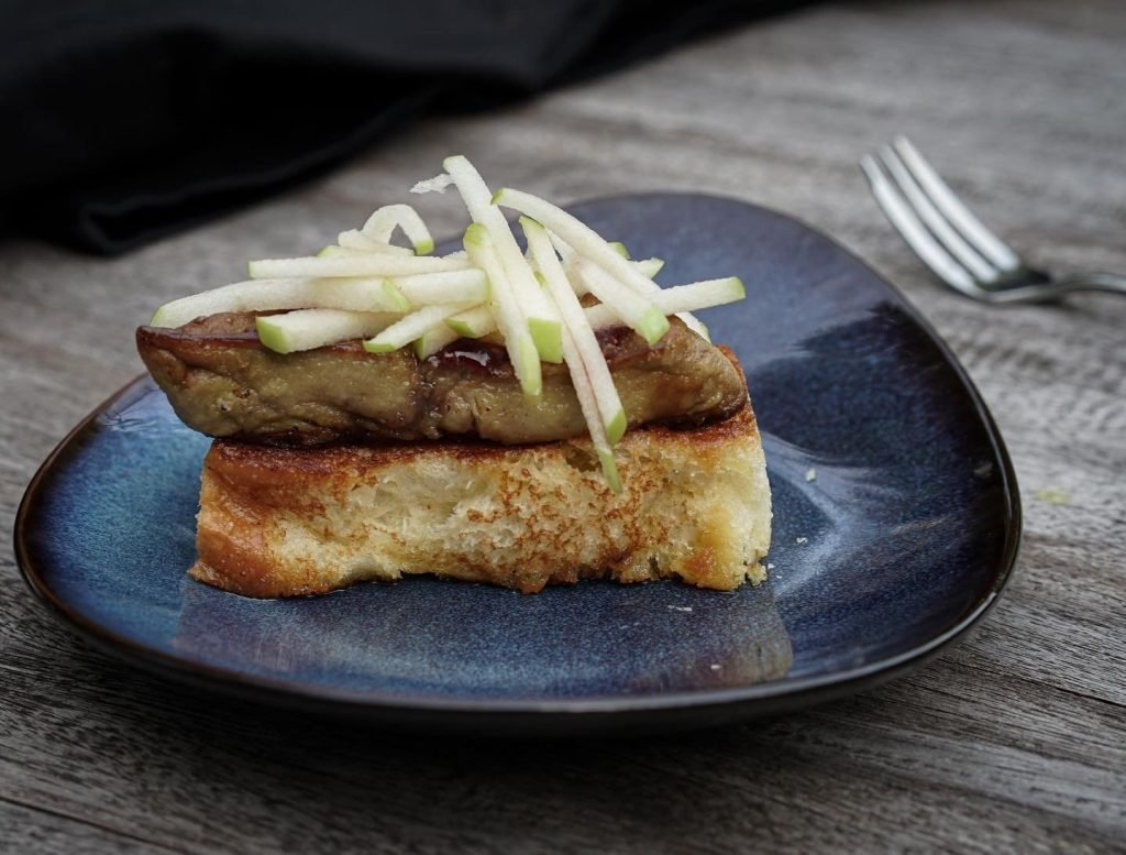 An elegant appetizer from CM-PB Contemporary Melting-pot, featuring a slice of seared foie gras on a thick piece of toasted brioche, topped with julienned green apple