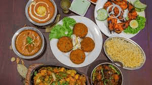 Overhead view of a colorful spread of popular Indian dishes on a dark tabletop, including tandoori chicken, various curries, kebabs, rice, and a sizzling platter.