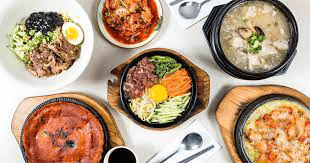 Flat lay of various Korean dishes including a central bowl of bibimbap with egg yolk, kimchi, samgyetang (chicken soup), kimchi jeon (pancake), and a cheesy baked dish.