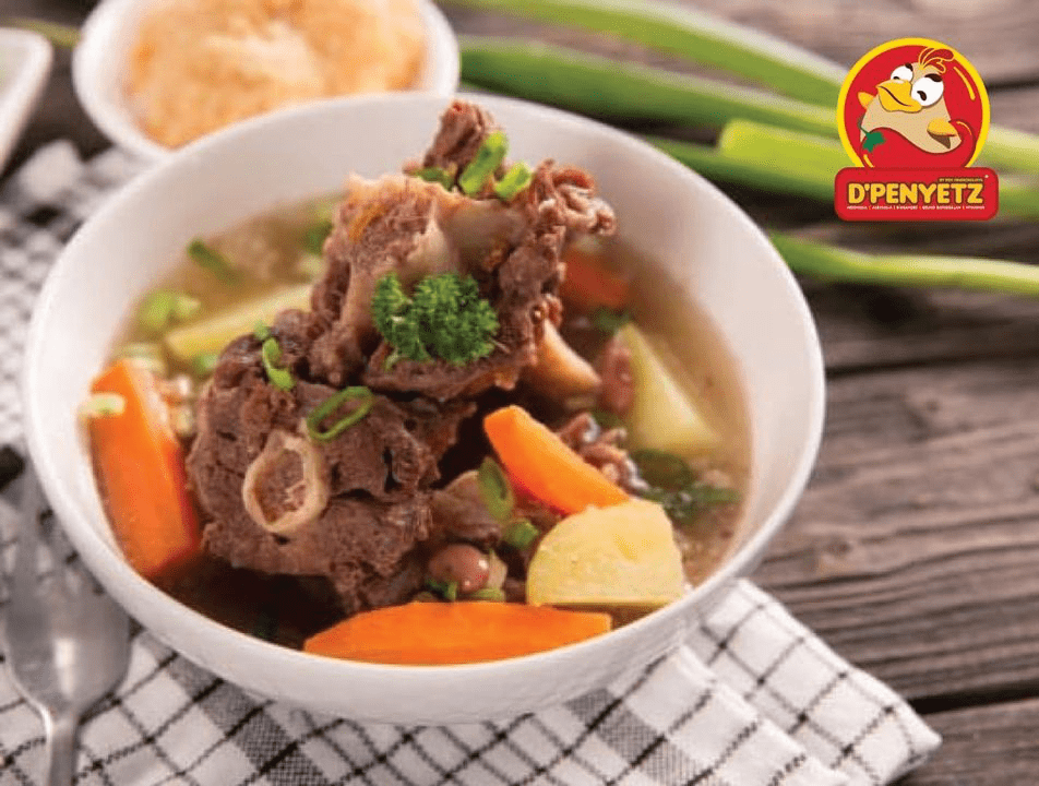 D'PENYETZ Oxtail Soup in a white bowl, featuring tender oxtail pieces, carrots, potatoes, and spring onions