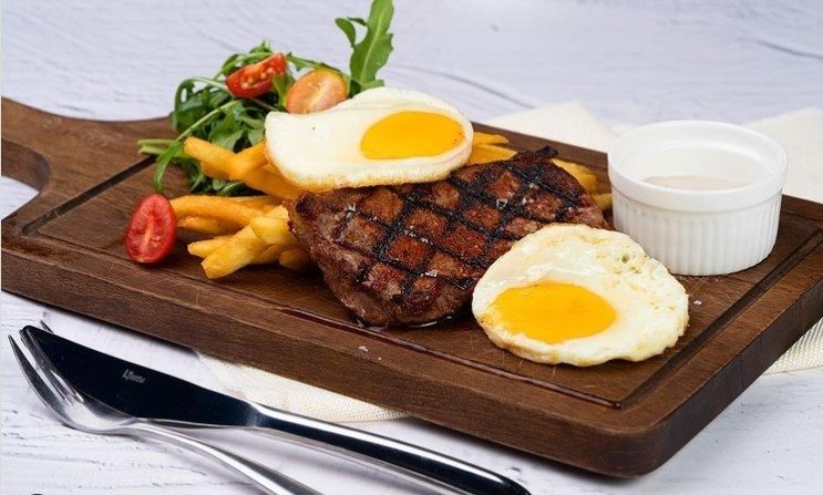 A meal from Drinks & Co. Kitchen served on a dark wooden board, featuring a grilled steak with prominent crosshatch grill marks, topped with one fried egg and another alongside, accompanied by French fries, a fresh arugula and cherry tomato salad, and a small white ramekin of sauce.