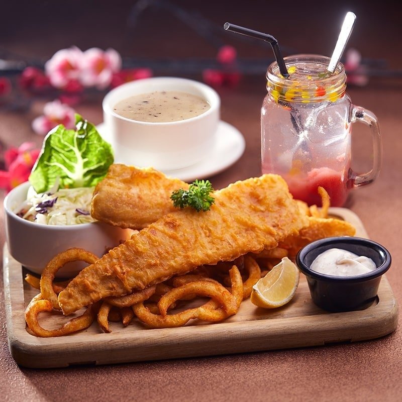 A meal set from Eighteen Chefs The Star Vista, featuring a large piece of golden-fried fish on a bed of curly fries, served on a wooden board with a side of coleslaw, tartar sauce, a lemon wedge