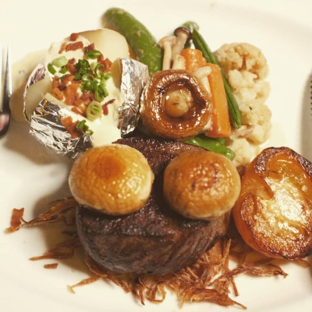 Close-up of a plated meal from Fosters Restaurant featuring a grilled steak topped with two button mushrooms, resting on a bed of crispy fried onions.