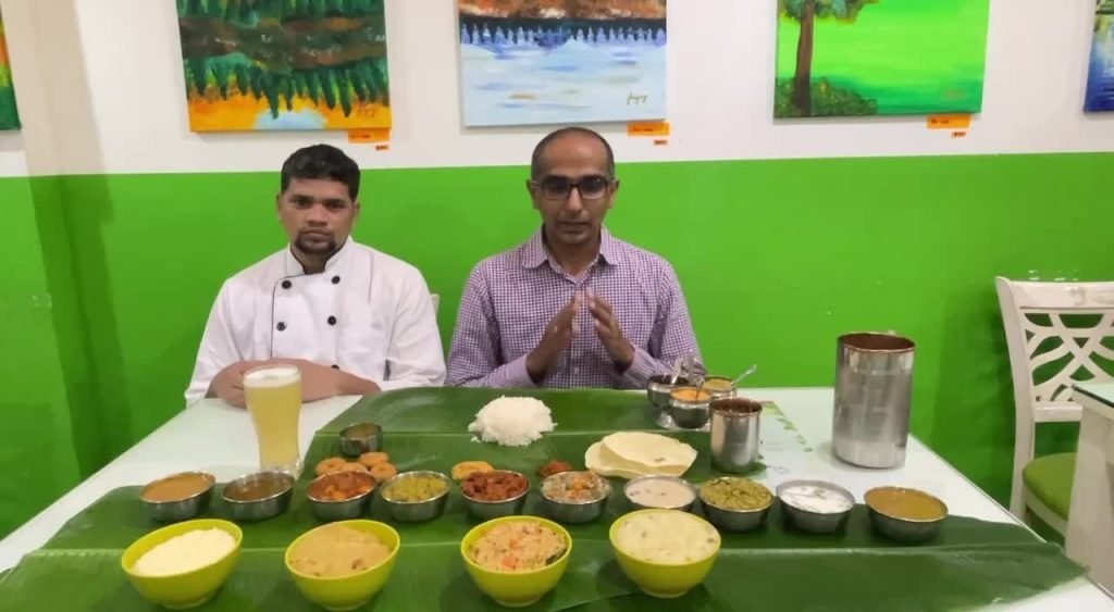 Two men at Greenleaf Cafe, one a chef and the other likely a host, presenting an elaborate traditional South Indian meal