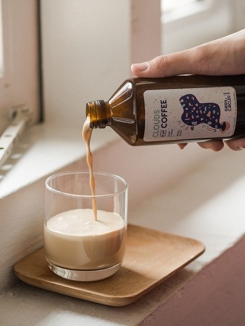 A hand pouring 'Clouds Coffee' cold brew from Grids & Circles from an amber glass bottle with a distinctive label into a clear glass