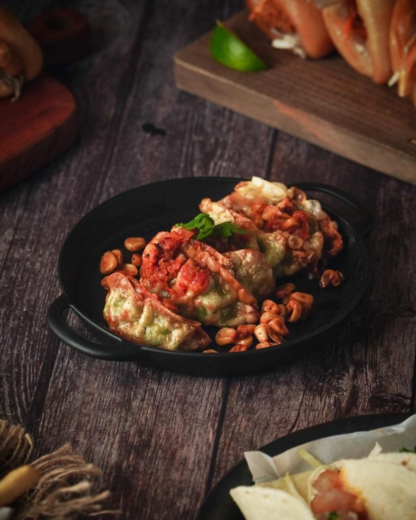 A dish from Harry's Holland featuring pan-fried green dumplings topped with a spicy red relish and cilantro, served with roasted broad beans in a black cast-iron style skillet on a dark wooden table.