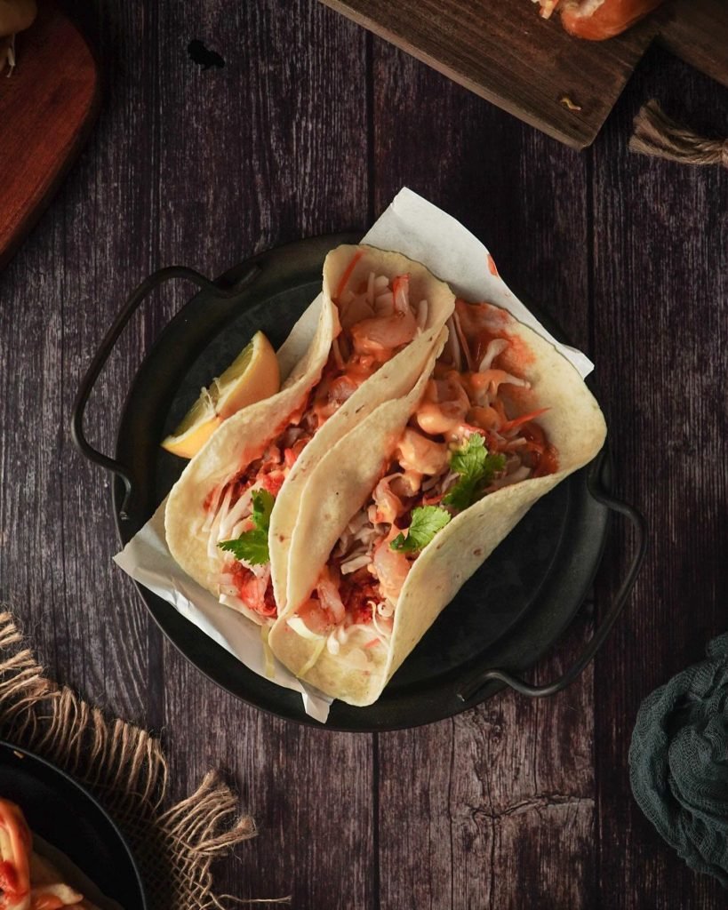 Three soft tacos from Harry's Holland, generously filled with shredded slaw, a creamy sauce, and cilantro, presented on a dark metal tray lined with paper, accompanied by a lime wedge on a dark wooden table.