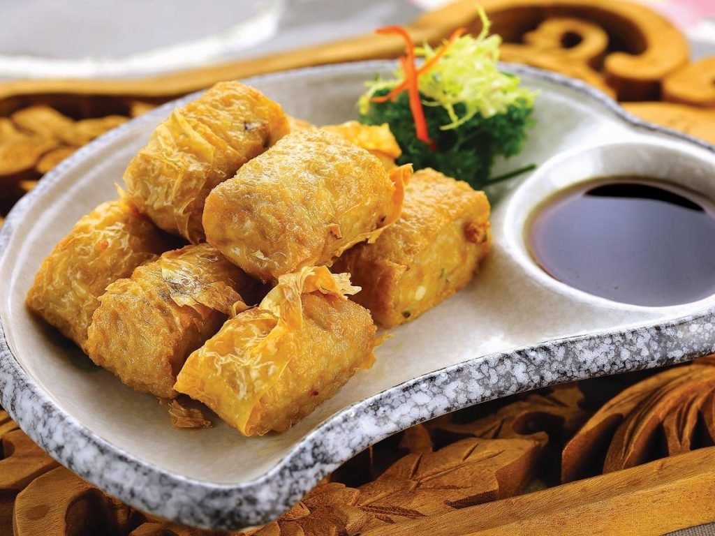 Golden fried beancurd skin rolls from Hilltop Garden Restaurant, neatly arranged on a speckled ceramic platter with an integrated bowl of dark dipping sauce and a fresh green garnish