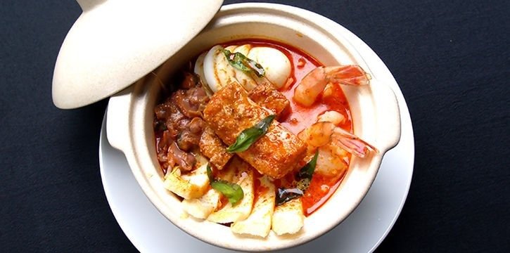 Claypot laksa from Hooha Cafe, featuring prawns, cockles, tofu puffs, hard-boiled egg, and rice cakes in a rich, reddish-orange curry broth, served in a light-colored clay pot with the lid ajar on a white saucer against a black background