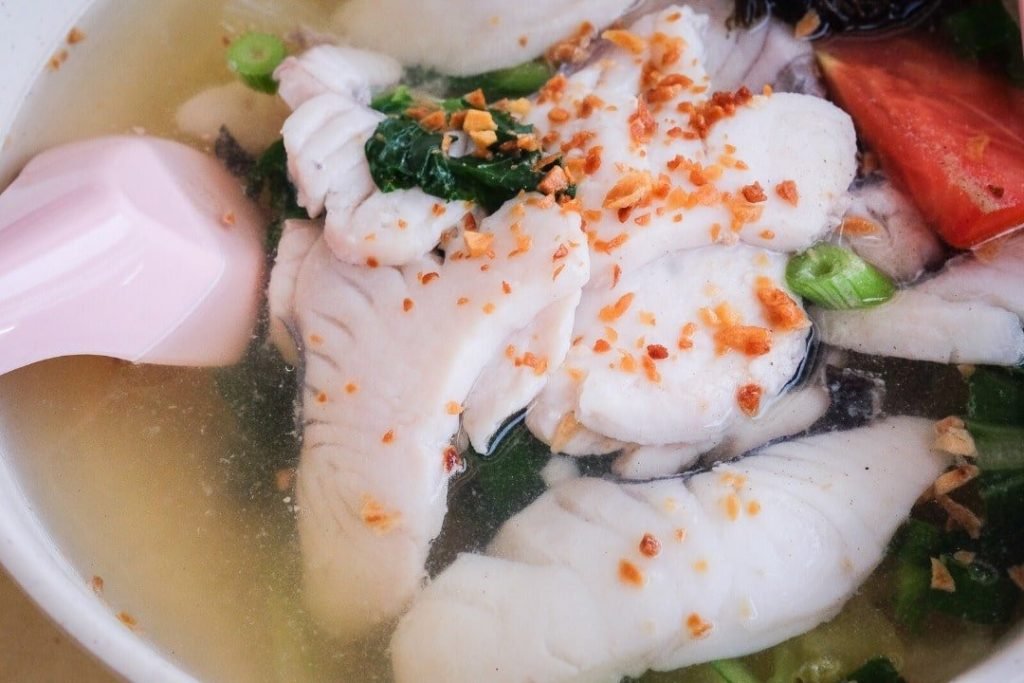 Close-up of sliced fish soup from Hougang Lee Chen Cooked Food, featuring tender white fish slices in a clear broth with green vegetables, a wedge of tomato, and topped with crispy fried garlic, served in a white bowl with a pink plastic soup spoon.