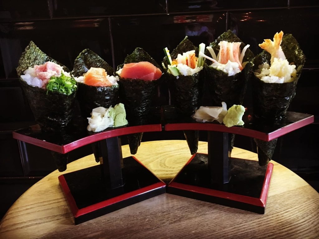 An assortment of six temaki hand rolls from IKKI IZAKAYA, including tuna, salmon, negitoro, crab stick, and shrimp tempura, presented on two black and red tiered stands with wasabi and pickled ginger on a wooden table.