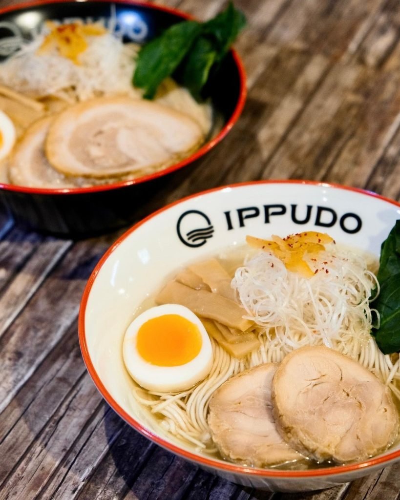 Two bowls of IPPUDO ramen on a wooden table, with the foreground bowl clearly displaying chashu pork, a soft-boiled egg, bamboo shoots, shredded leeks, and noodles in broth, with the IPPUDO logo visible on the bowl