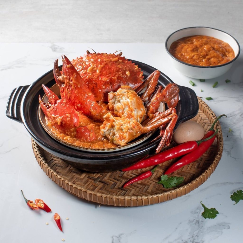 Jiak Modern Tzechar's signature Chilli Crab dish, presented in a black claypot on a woven rattan tray, accompanied by a side bowl of extra sauce, fresh red chilies, an egg, and cilantro sprigs on a white marble surface