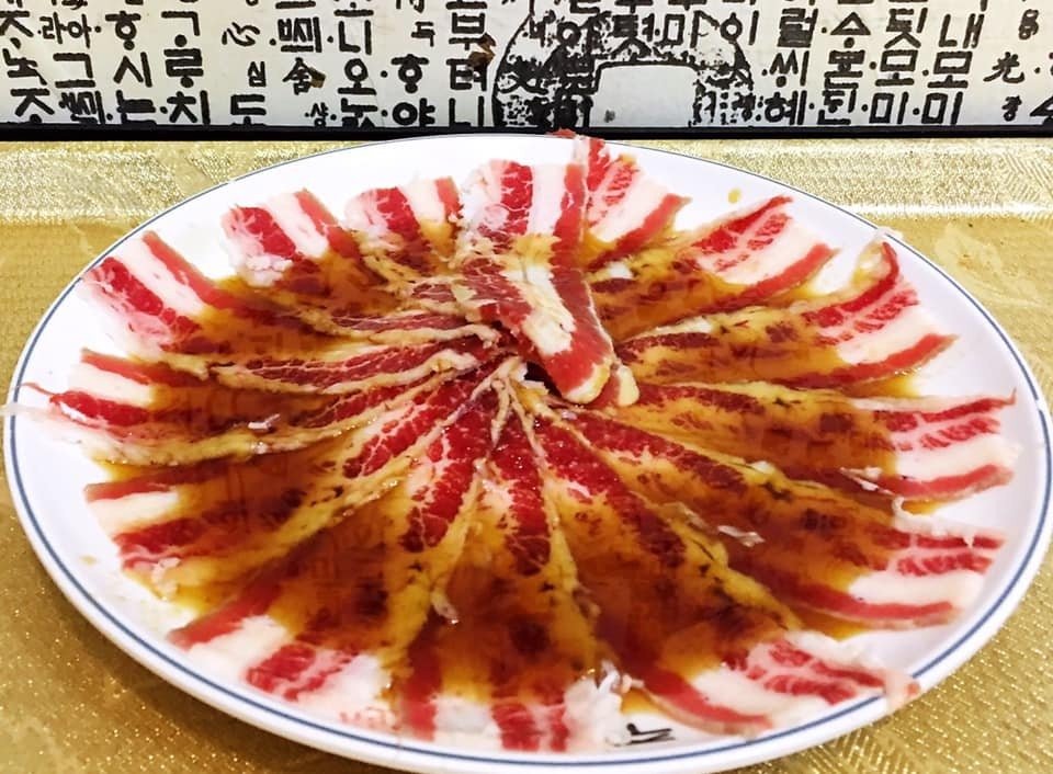 A white plate from Ju Shin Jung featuring thinly sliced raw beef, possibly brisket, fanned out in a circular pattern and drizzled with a dark marinade, ready for Korean BBQ, with Korean script visible in the background.
