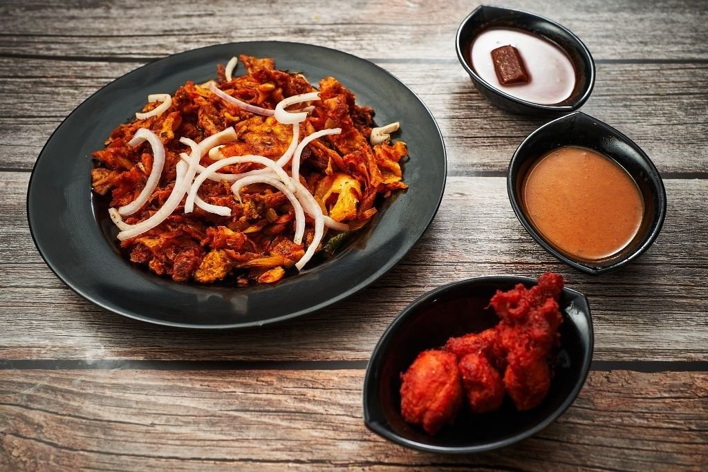 A meal from Kings Indian Banana Leaf Restaurant featuring a plate of kothu parotta topped with onion rings, a side of red fried chicken pieces, and two dipping sauces, all arranged on a wooden table.