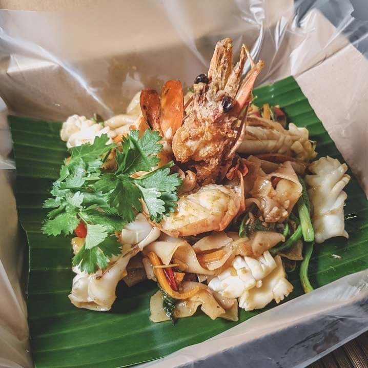 Close-up of a Thai seafood noodle dish from Kra Pow Thai Restaurant, featuring a large fried prawn, squid, flat rice noodles, and cilantro, served on a banana leaf
