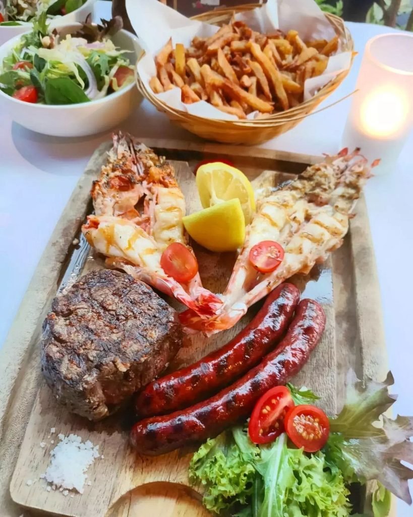 A hearty mixed grill platter from Les Bouchons Rive Gauche, including a beef steak, grilled prawns, and sausages, presented on a wooden board with sides of French fries and salad, in a candlelit setting.