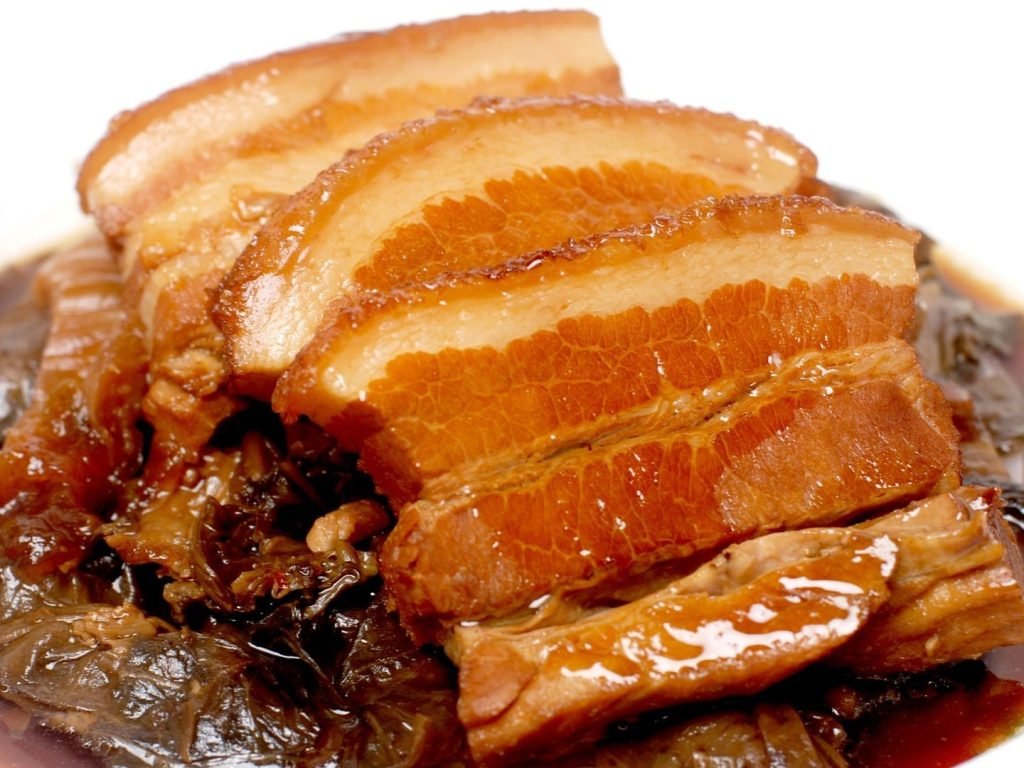 Close-up of tender, glistening slices of braised pork belly served over dark preserved mustard greens