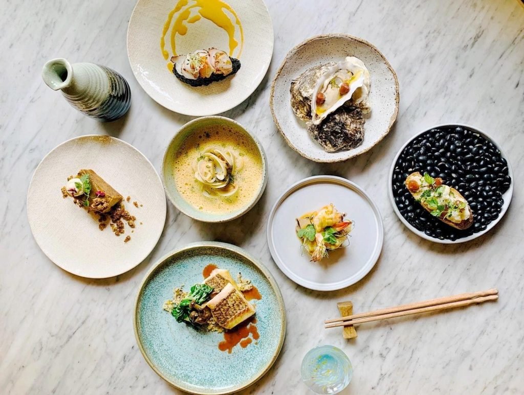 An artistic spread of various Asian-fusion small plates from Morsels restaurant, including seafood dishes like fish, oyster, and clams, a terrine, and a prawn item, arranged on a marble table with a sake flask and chopsticks