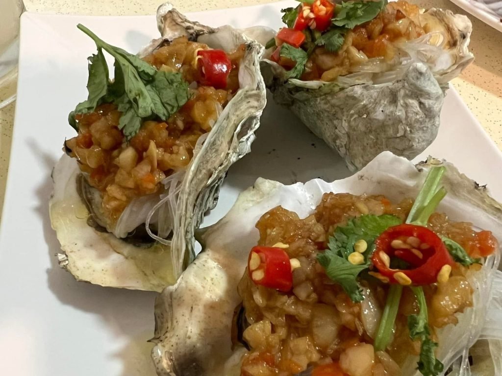 A plate of three Chinese-style steamed oysters from Oriental Chinese Restaurant, each topped with a flavorful mince of garlic and chili, served on a bed of glass noodles and garnished with fresh cilantro and red chili.