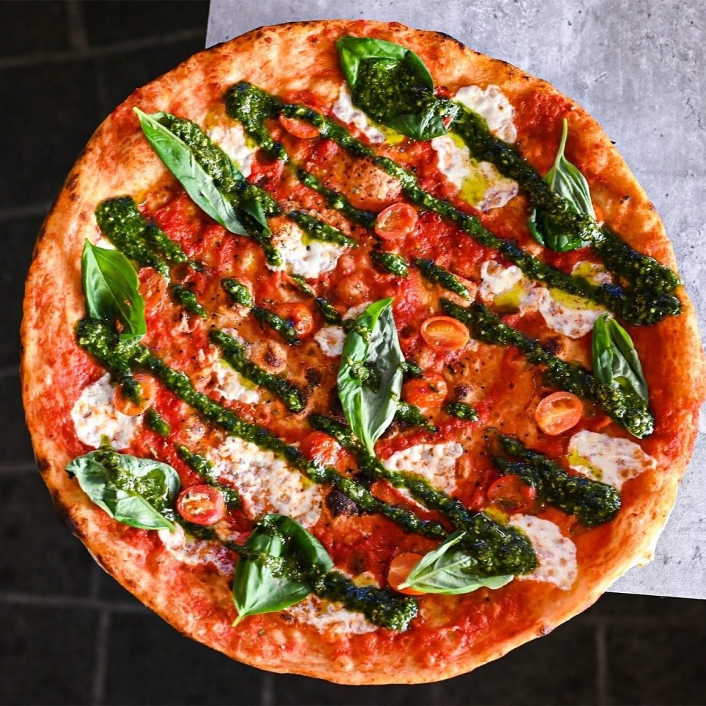 Overhead shot of a delicious Pizza Express pizza topped with tomato sauce, fresh mozzarella, green pesto drizzle, fresh basil, and cherry tomatoes.