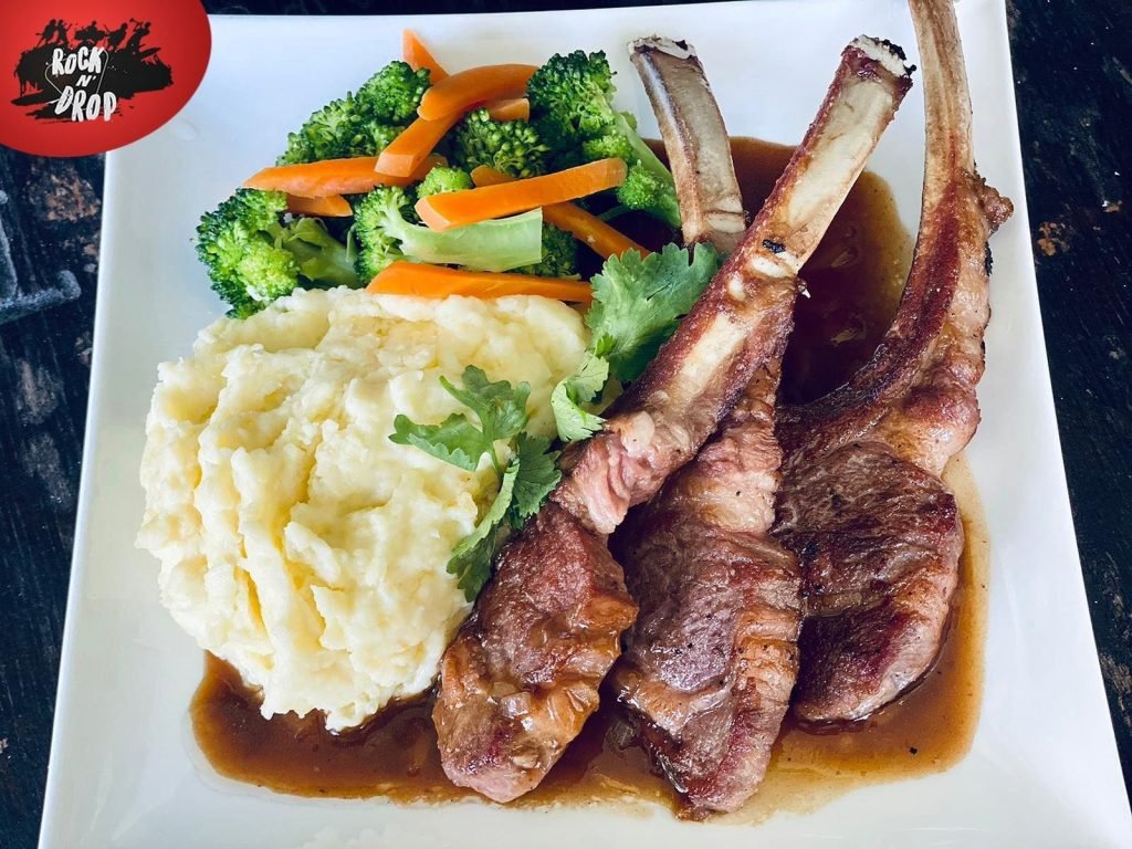 Plated meal from Rock N' Drop: Three frenched lamb chops with gravy, served with mashed potatoes, steamed broccoli, and carrots, and garnished with cilantro