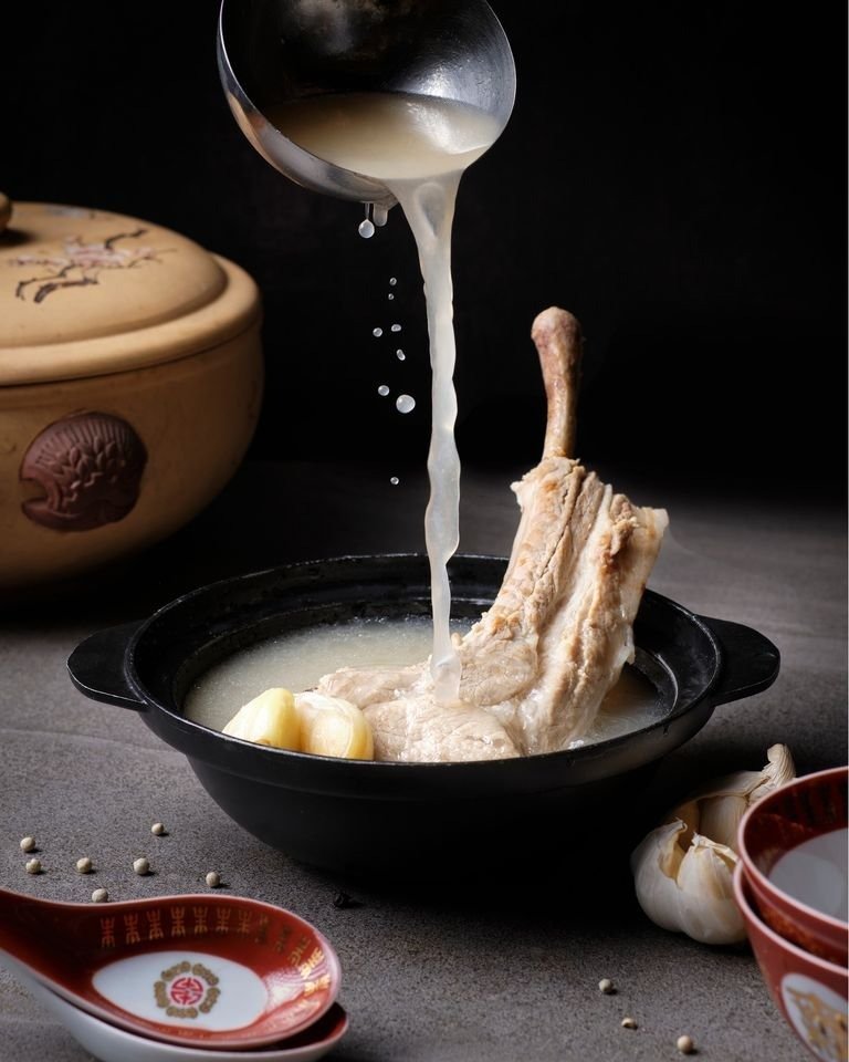 A ladle pours hot, milky broth over tender pork ribs and large garlic cloves in a black serving bowl of Bak Kut Teh