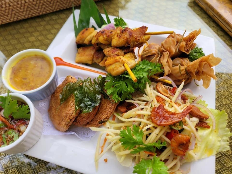 A Thai appetizer platter from Royal Thai Boat Noodles & Bar, featuring chicken satay, fried money bags, fish cakes, and green mango salad, served with two dipping sauces