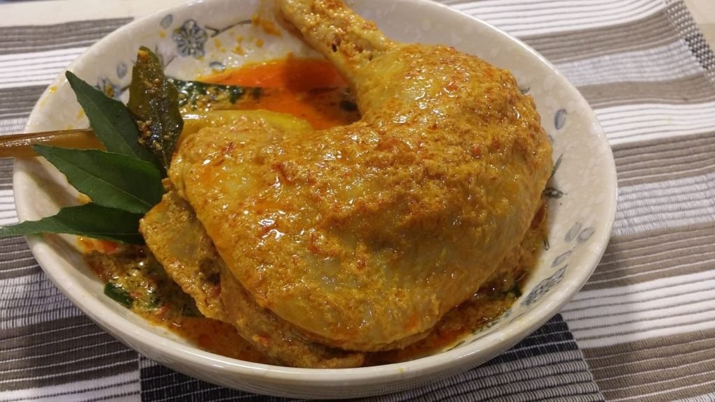 Close-up of a homemade chicken curry featuring a whole chicken leg quarter smothered in a savory orange sauce, garnished with curry leaves