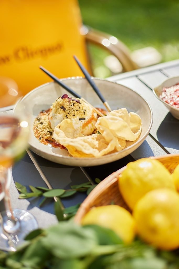 An elegantly presented dish in a ceramic bowl, possibly burrata or a savory ice cream, topped with crumbles and a red drizzle, served with crispy papadum-like chips and black chopsticks, on an outdoor slatted table
