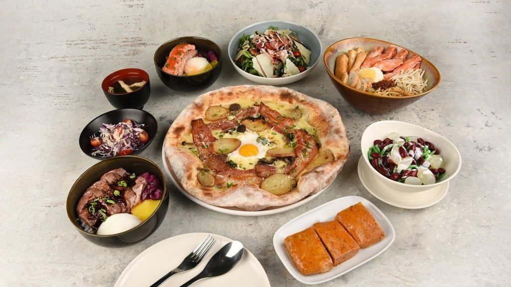 A large spread of diverse dishes from SOCIEATY, featuring a central pizza with egg and bacon, surrounded by bowls of prawn noodles, beef donburi, salmon donburi, salads